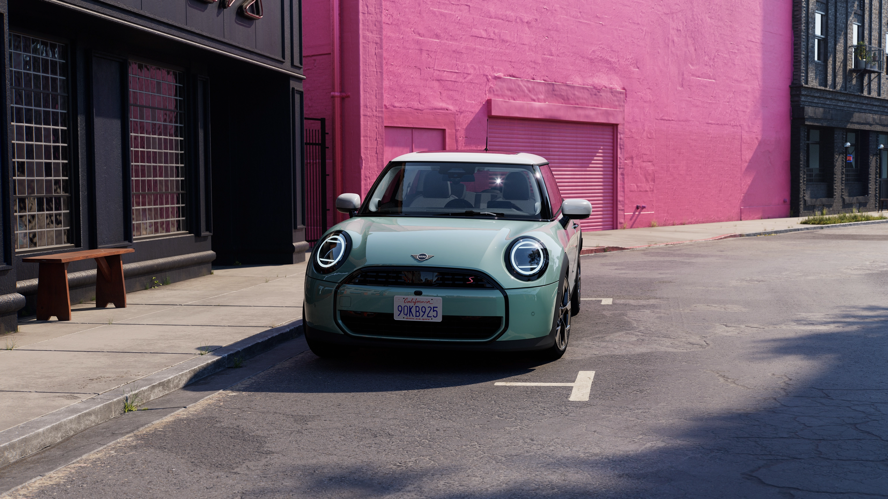 Prednji prikaz MINI Cooper-a Ocean Wave zelene boje sa belim krovom dok se parkira na ulici sa ružičastim zidom u pozadini