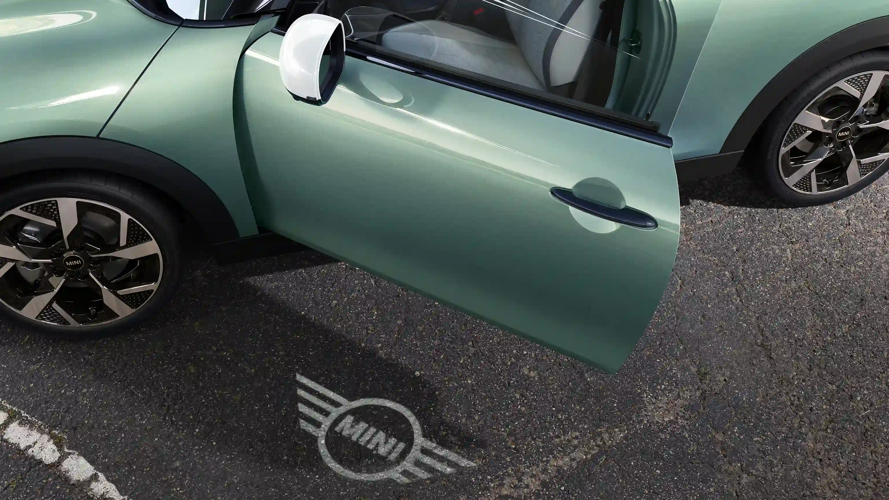Spoljašnji prikaz MINI Cooper-a sa otvorenim vratima i MINI svetlom dobrodošlice sa projekcijom logotipa na zemlji 