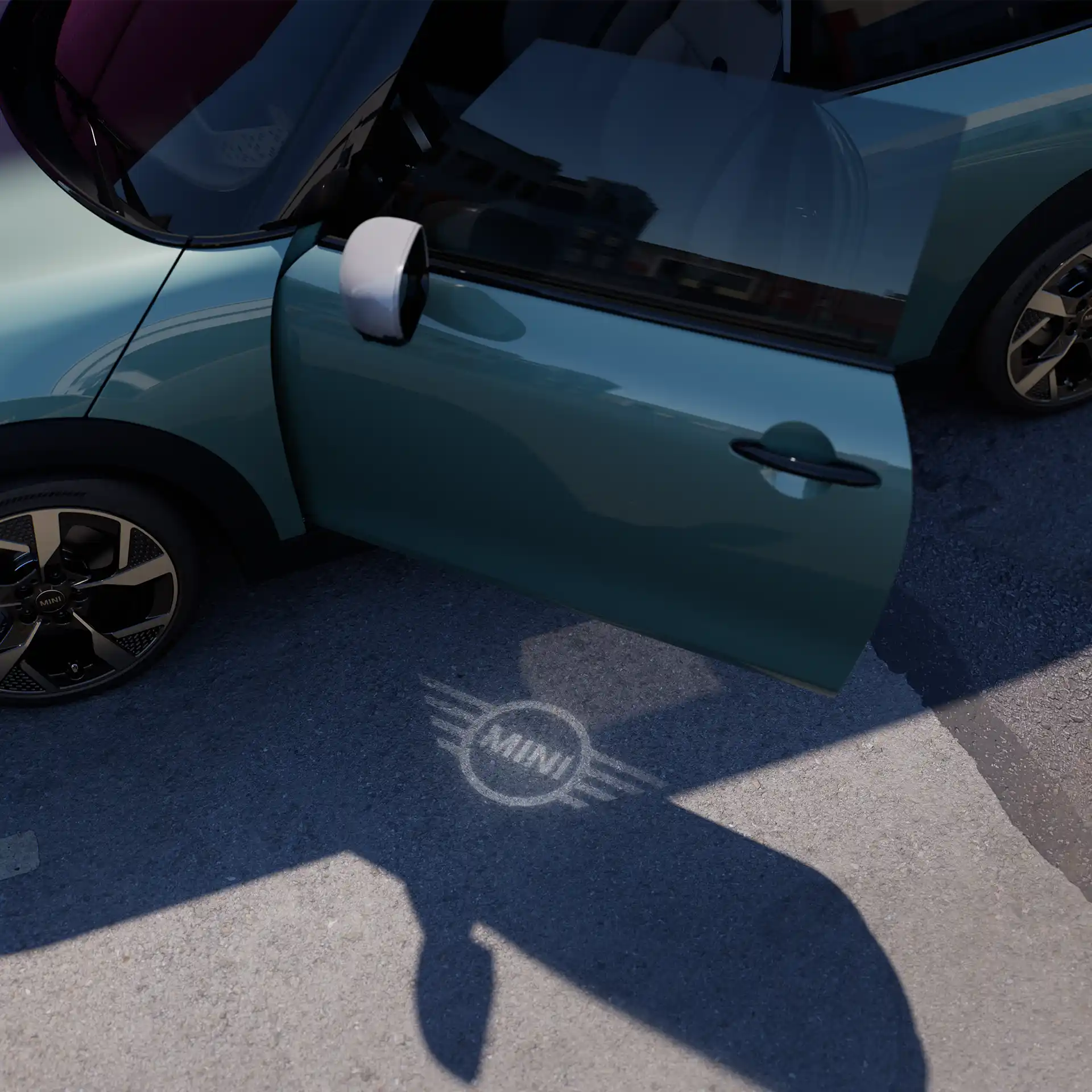 Spoljašnji prikaz MINI Cooper-a sa otvorenim vratima i MINI svetlom dobrodošlice sa projekcijom logotipa na zemlji 