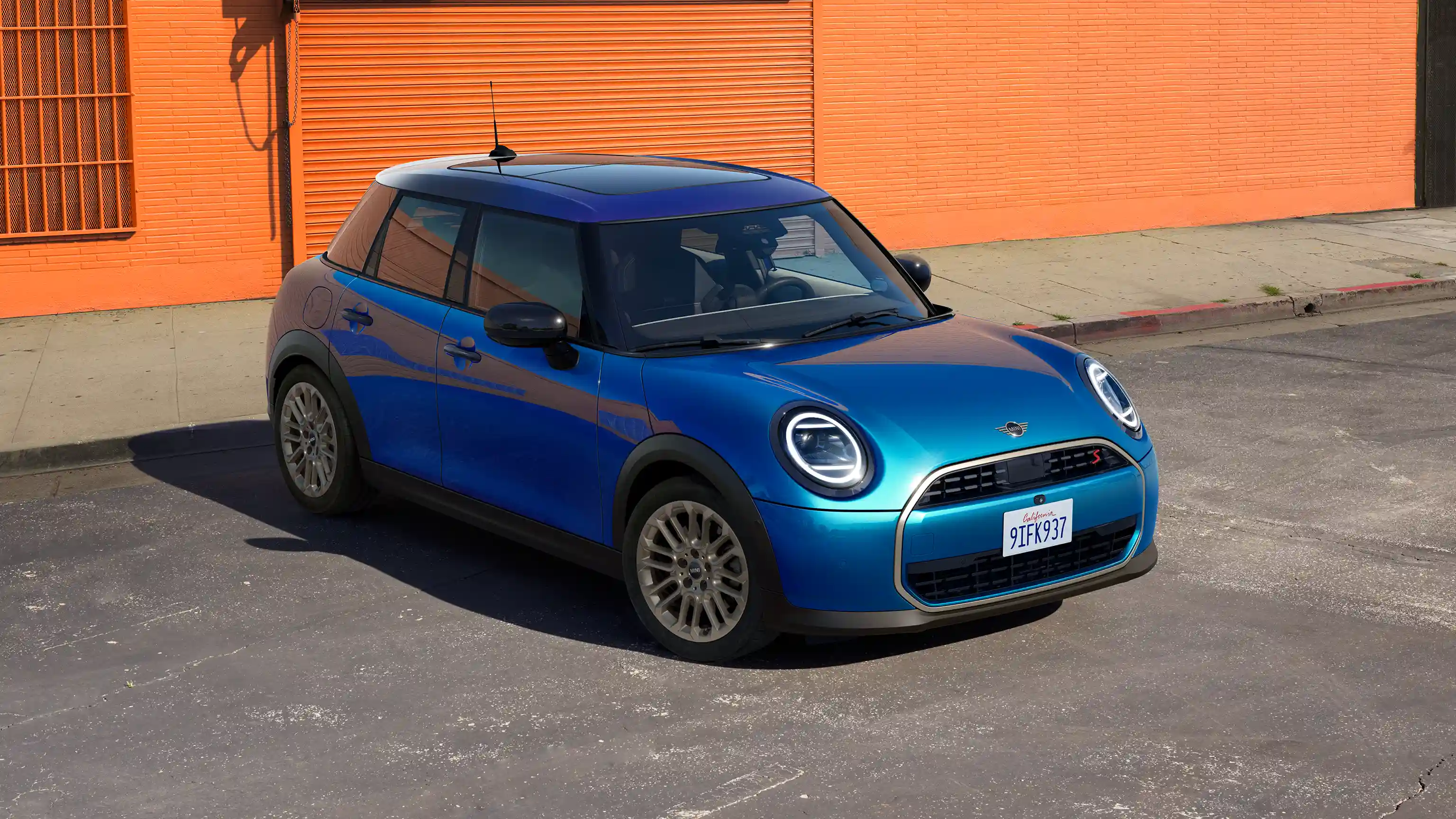 Prednji prikaz MINI Cooper-a sa 5 vrata Icy Sunshine plave boje sa višenijansnim plavim krovom prilikom parkiranja na ulici sa narandžastim zidom u pozadini