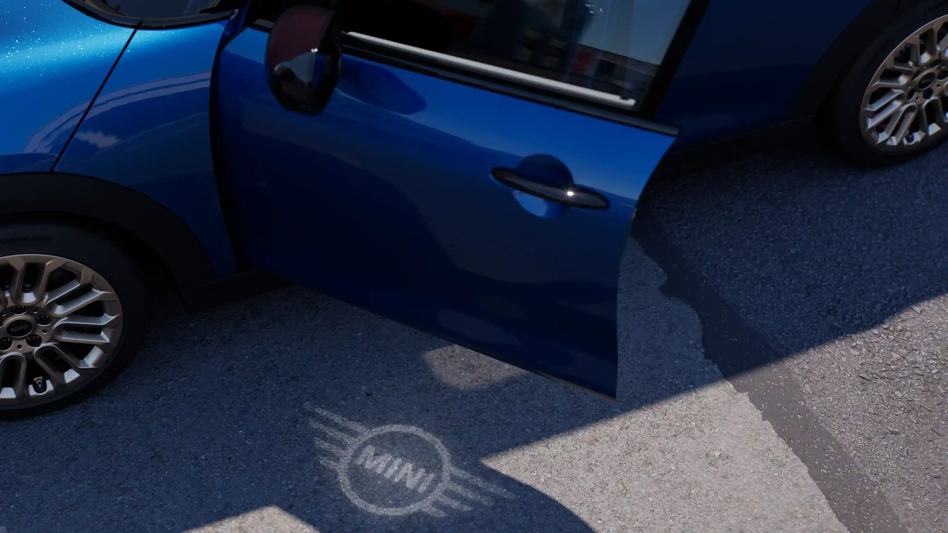 Spoljašnji prikaz otvorenih vrata MINI Cooper-a sa 5 vratima i MINI svetla dobrodošlice sa projekcijom logotipa na zemlji 
