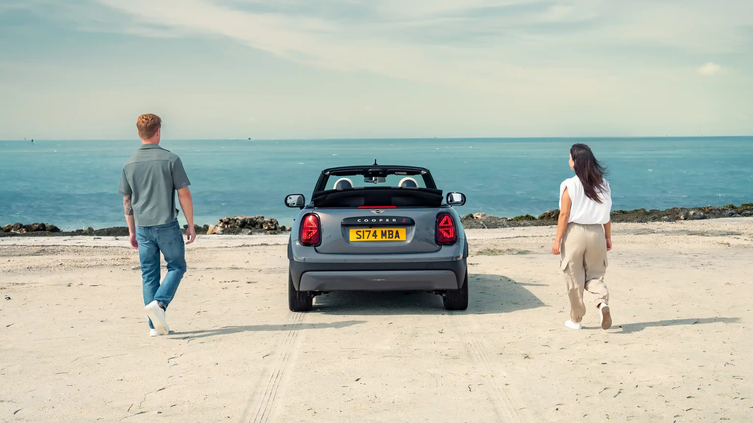 Prikaz zadnje strane parkiranog MINI Cooper Kabrioleta na plaži na kojoj se nalaze dve osobe