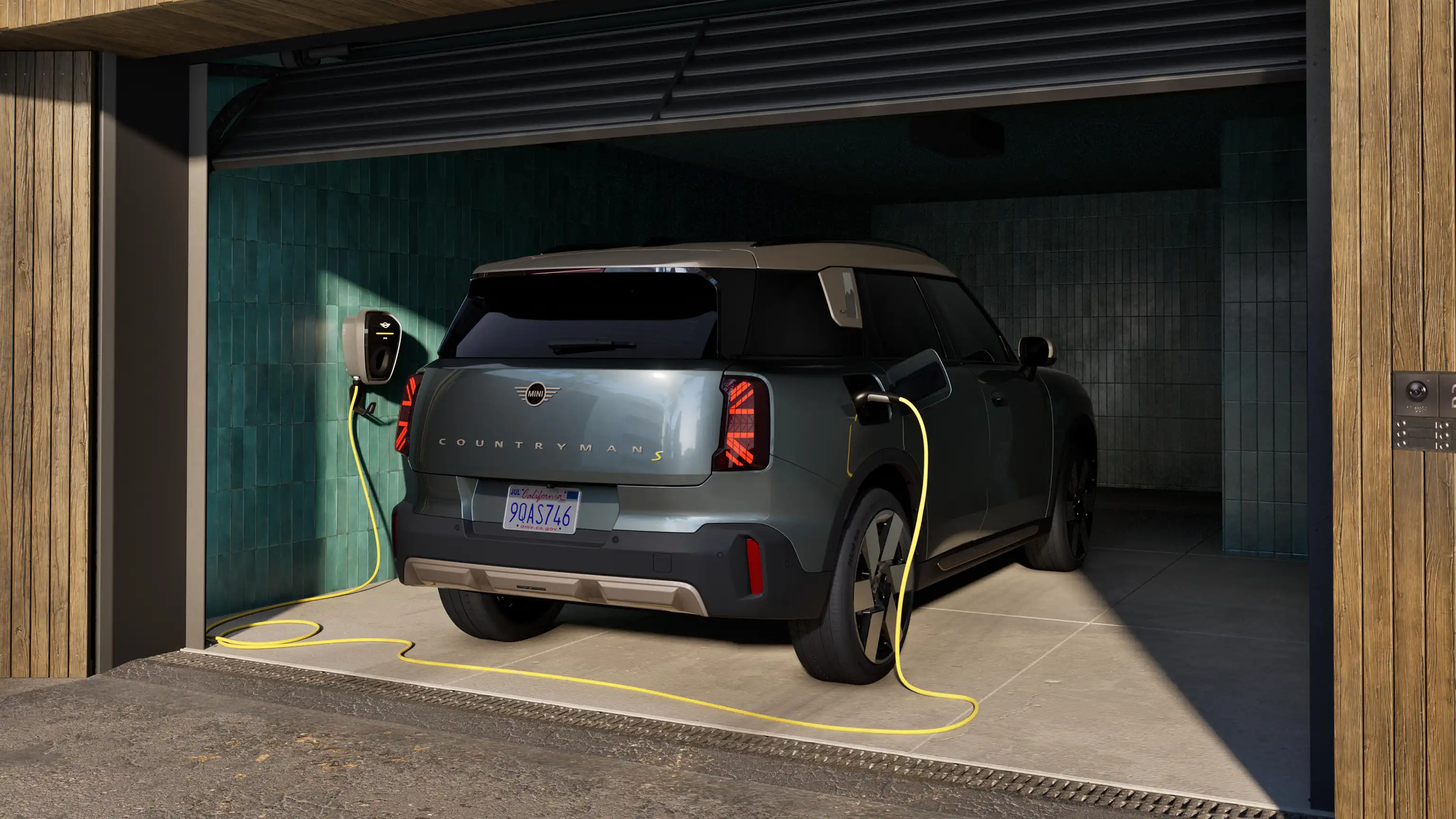 Prikaz zadnje strane potpuno električnog MINI Countryman-a Smokey zelene boje u otvorenoj garaži povezan kablom za punjenje.