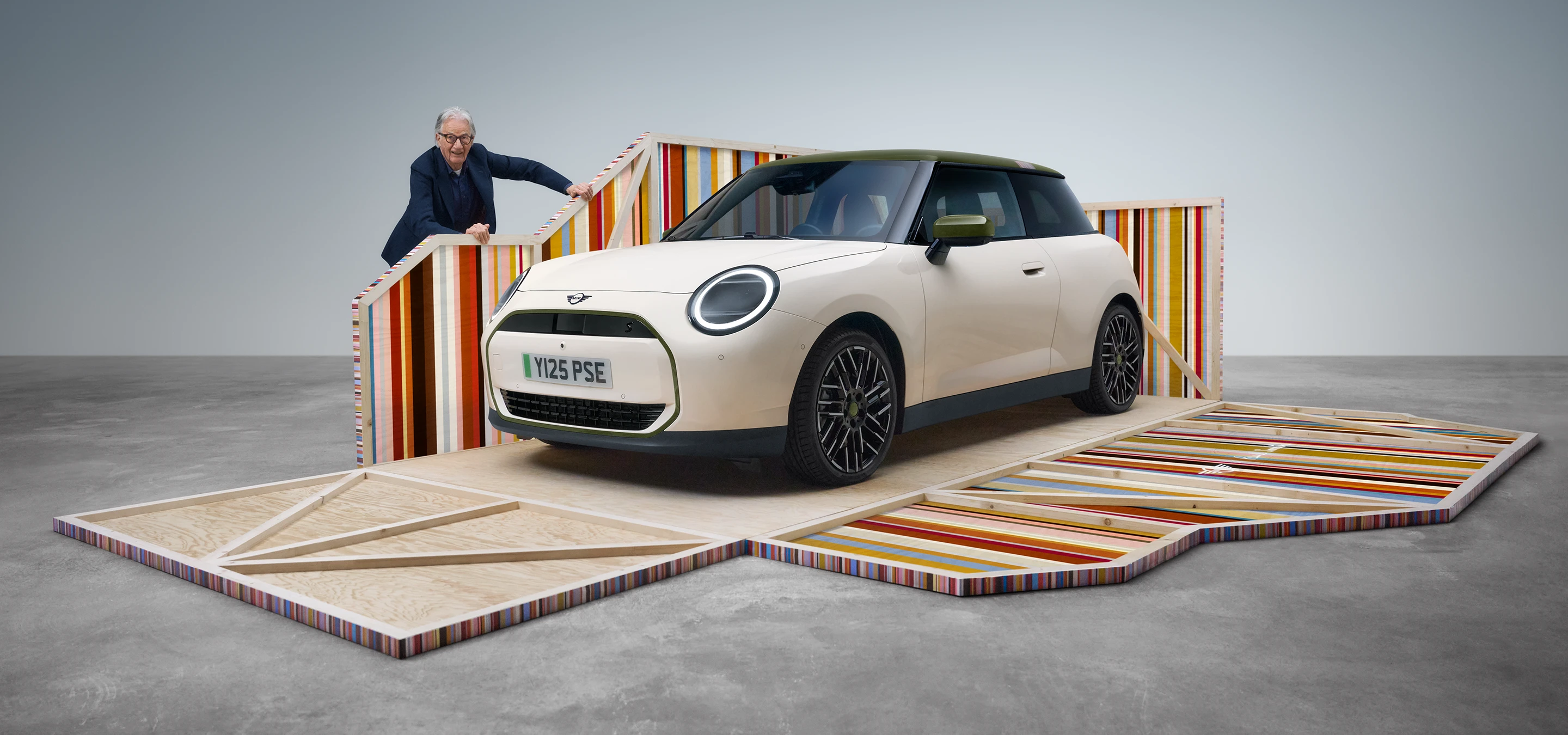 Pogled spreda sa strane na potpuno električni MINI Cooper Paul Smith izdanje u boji Inspired White, pored kog stoji dizajner Paul Smith.